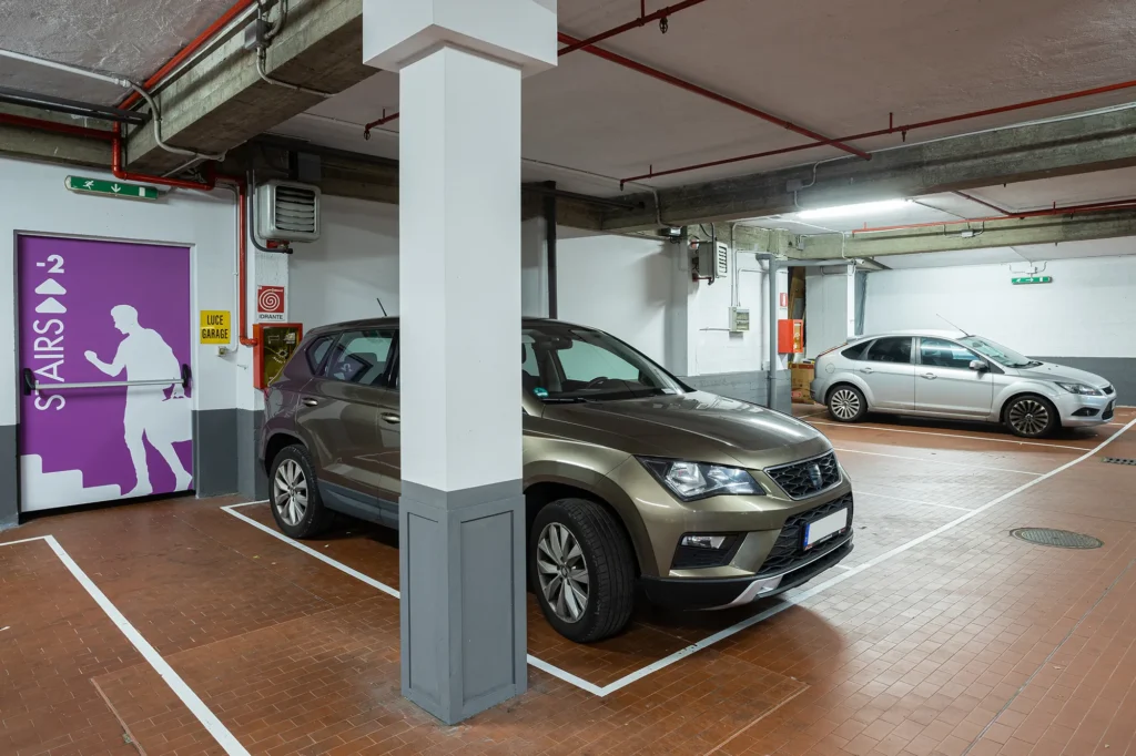 In the picture a detail of the hotel garage. You can see several parking spaces with a couple of cars parked. On the left a door with a sign for the stairs.