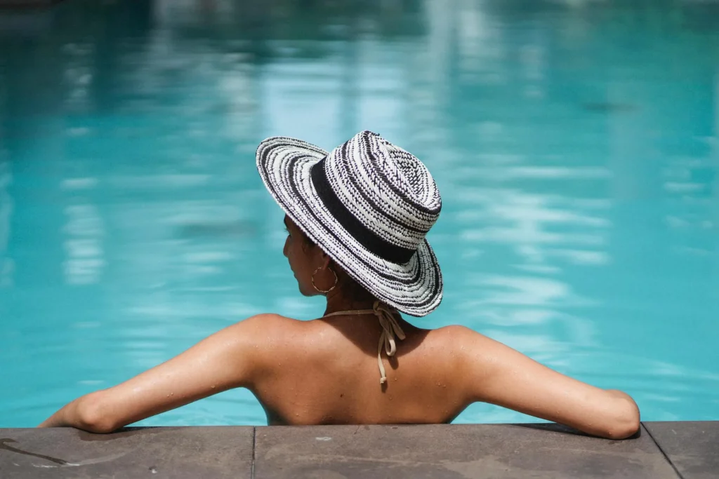 Una ragazza si trova all'interno di una piscina con la schiena e le braccia poggiate al bordo. Indossa un cappello di paglia rotondo nero e bianco e ha un laccio del bikini allacciato dietro il collo. Porta ai lobi degli orecchini a cerchio e guarda in lontananza